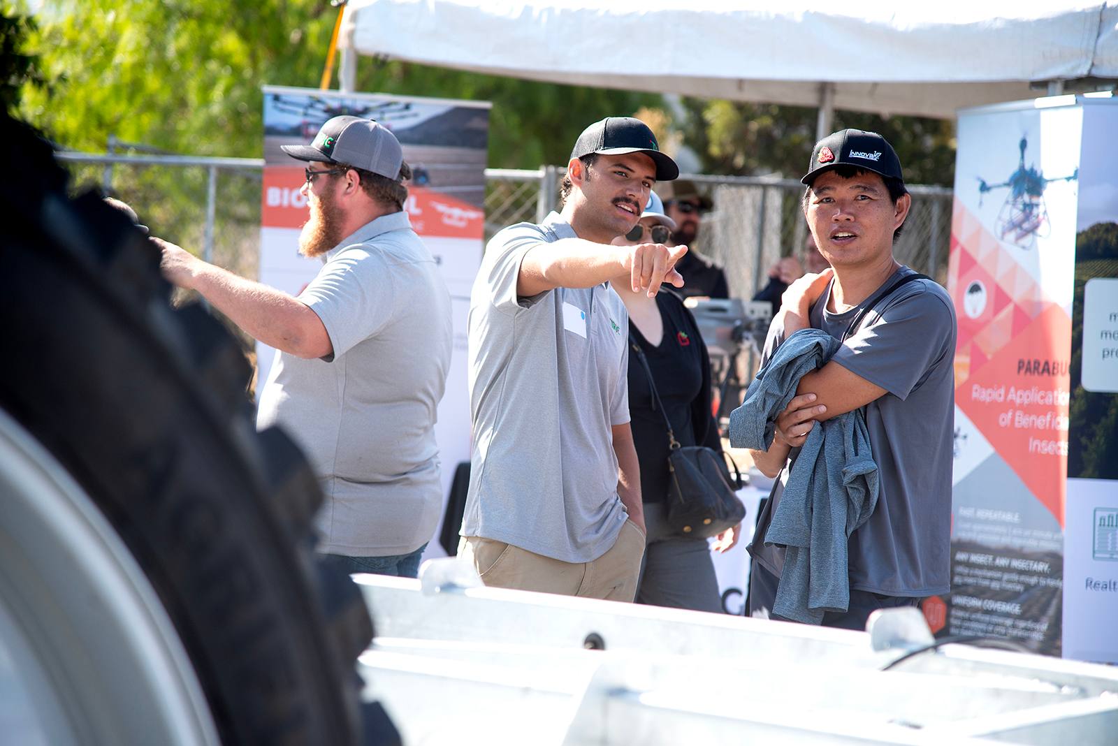 Two men at an outdoor agricultural technology expo discussing equipment, with a large tire and informational booths in the background.