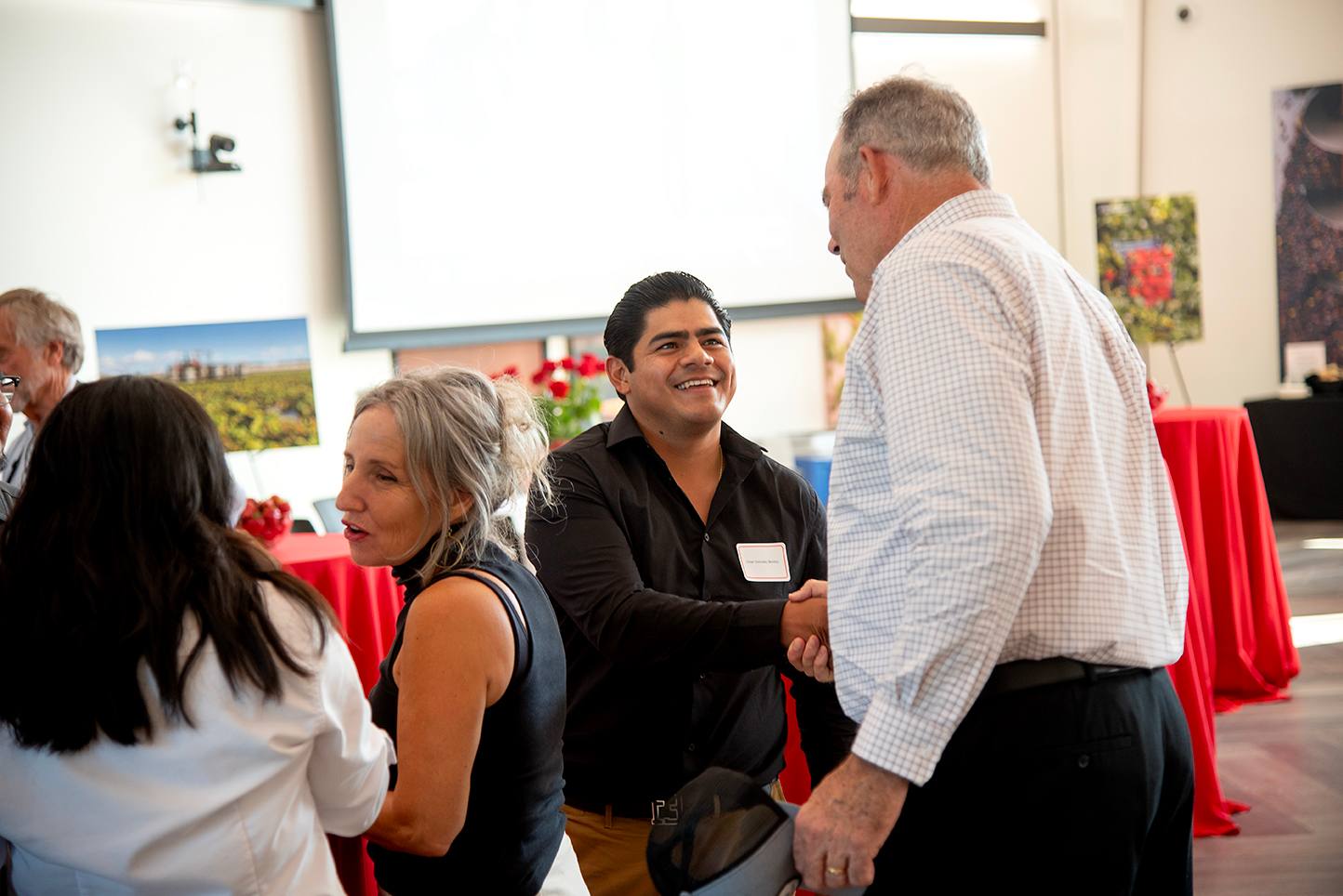 A smiling man shakes hands with another attendee during a networking event in a bright conference room decorated with red tablecloths and agricultural-themed photos.
