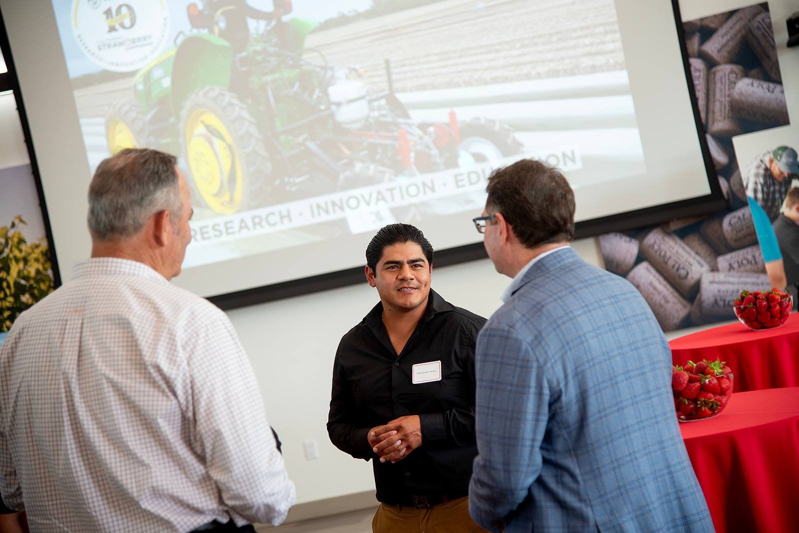 Man networking with two attendees at an agricultural innovation event, with a presentation slide showing a strawberry research project in the background.