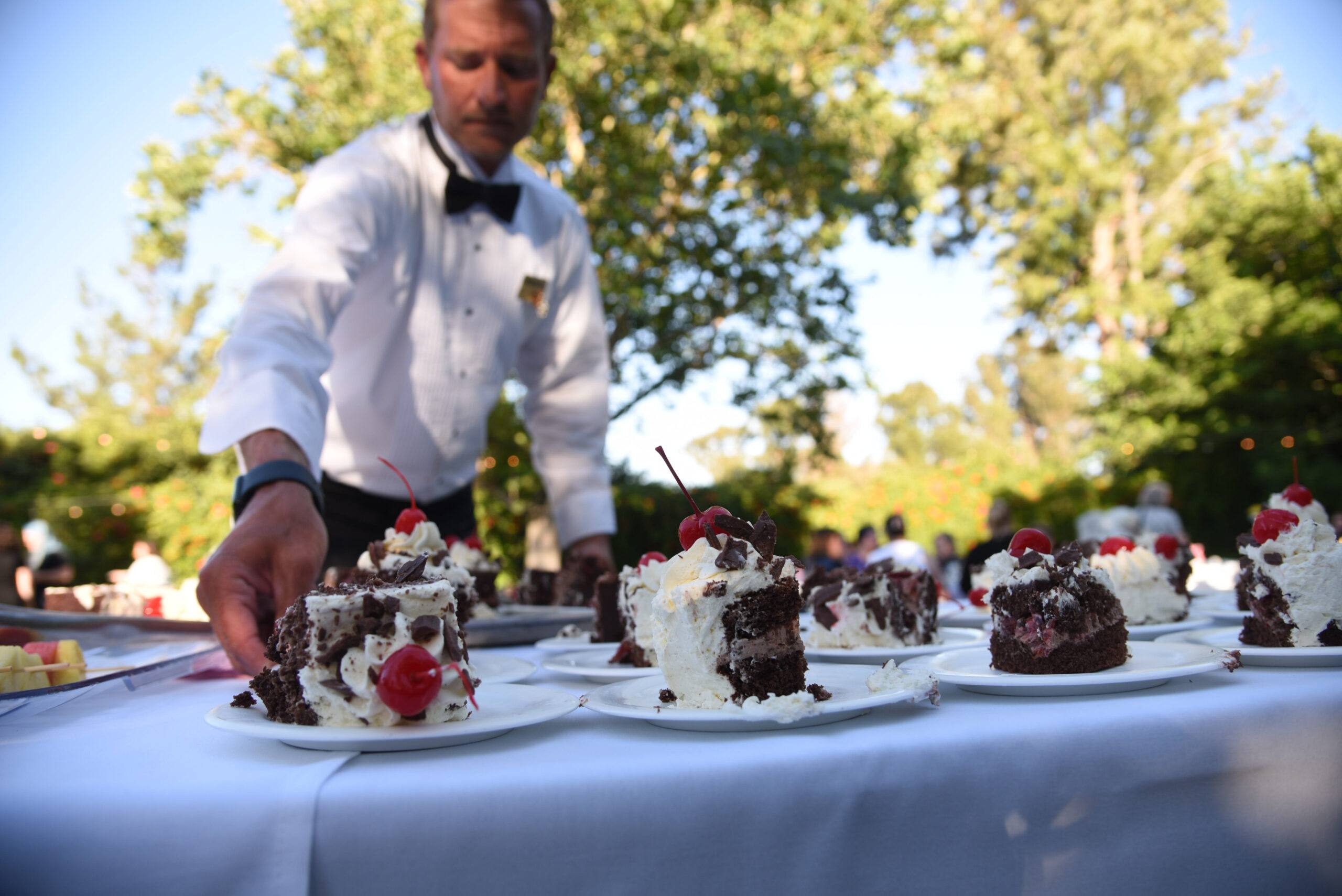 Waiter in formal attire serving plates of chocolate cake topped with whipped cream and cherries at an outdoor event.